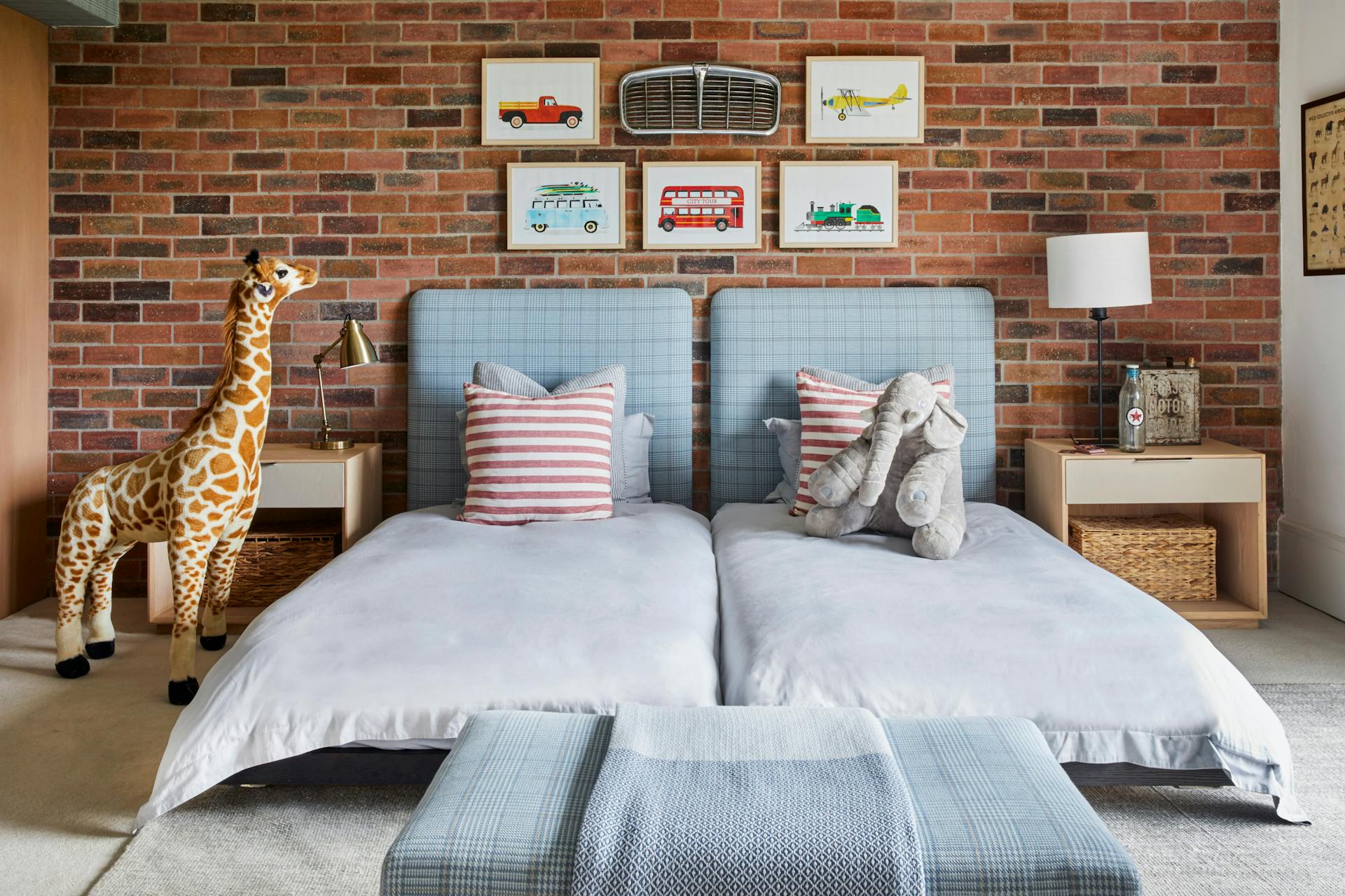 Two low-slung twin beds sit side by side in what appears to be a children's bedroom. The bed linen is a pale blue or grey, while the twin headboards and single ottoman are upholstered in plaid, and cushions on the bed are striped red. On one bed is a plush elephant, beside the other bed is a plush giraffe, and on the brick wall behind the bed are a vintage car's grille with five framed illustrations of vehicles: a pickup truck, small plane, VW van, double-decker bus and steam train.