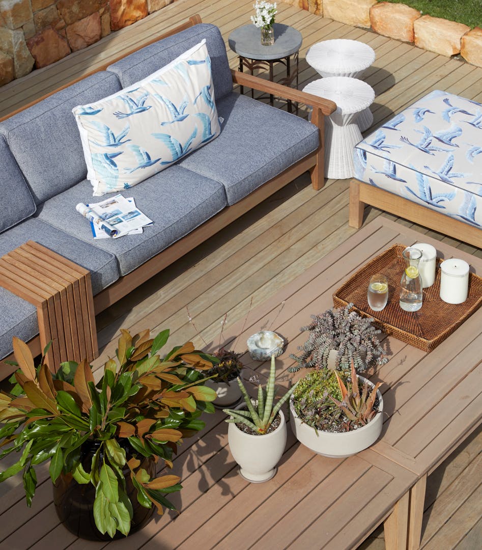 A collection of potted succulents stands on an outdoor table with a wooden appearance. Blue-upholstered outdoor furniture, a patterned ottoman and a collection of outdoor tables sit alongside it