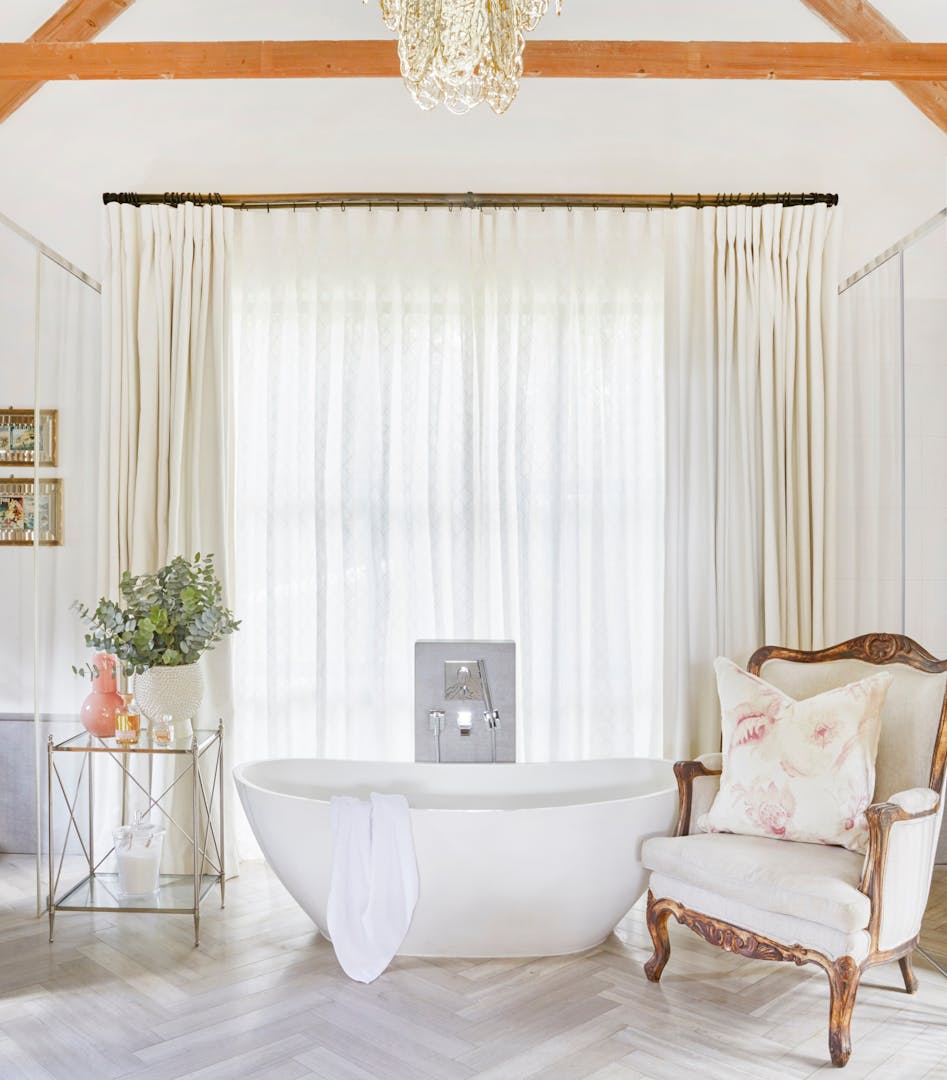A modern, freestanding bathtub is seen in the middle of a room with a white, bèrgere-style armchair to one side and a table, holding candles and a vase of greenery, on the other. There are sheer curtains behind the bath, and wooden beams and a glass chandelier above it.