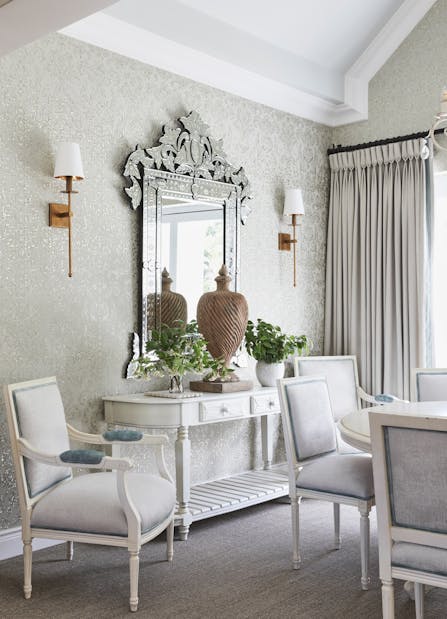 A white console table with decorative accents placed below an ornate mirror on the wall with a sconce positioned on each side.