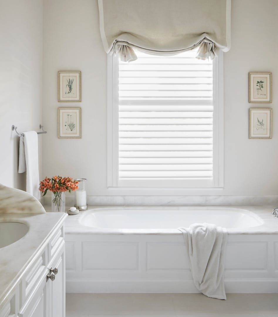 A bathroom in soft, white tones with a bathtub in the centre, frames on the wall and a basin to the left.