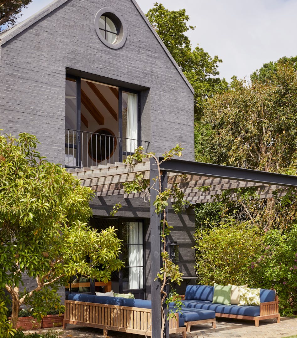 An A-frame, double-storey house painted in grey is surrounded by trees and other plant life. Wooden beams form a pergola-style cover for wooden, blue-upholstered outdoor furniture in an outdoor seating area