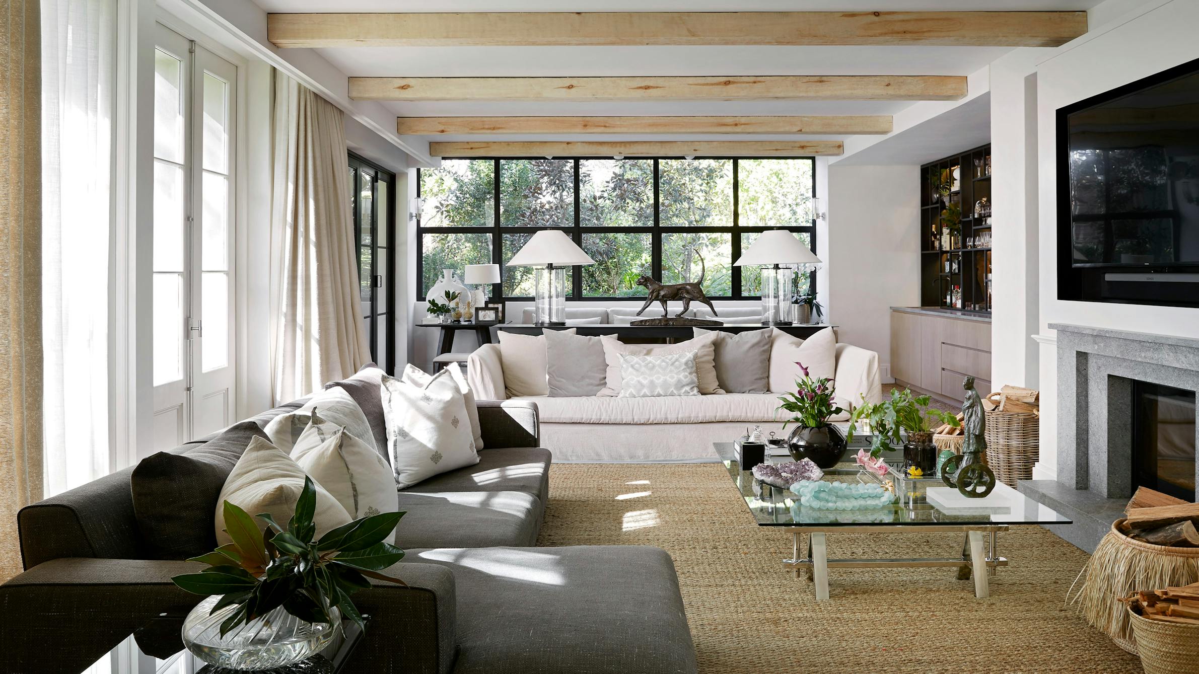 A contemporary living room with a glass coffee table in the centre, a grey and a white sofa on either side and large windows in the background.
