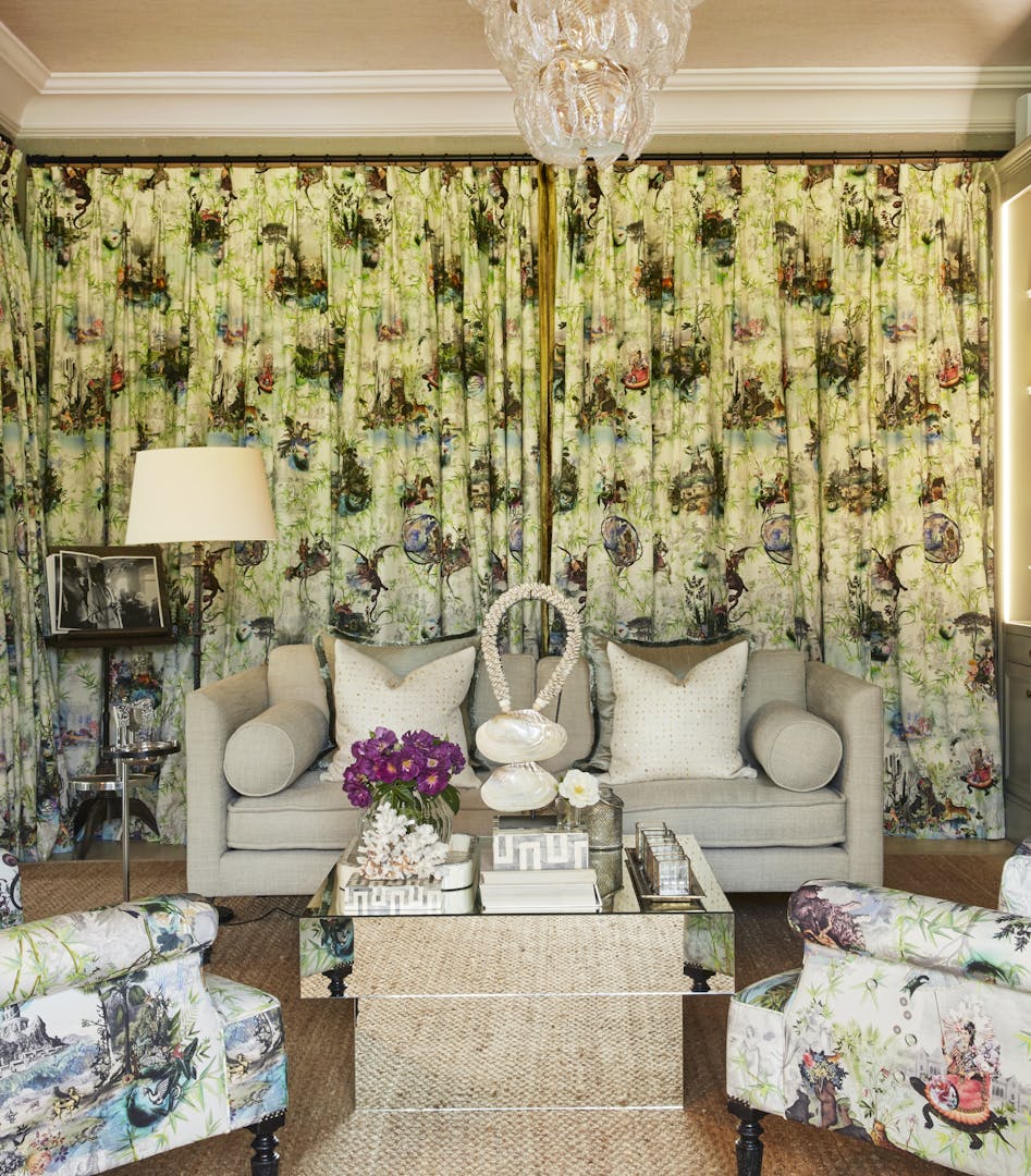A square, mirrored coffee table holds ornaments and flowers in front of a light-coloured sofa and intricately patterned curtains.