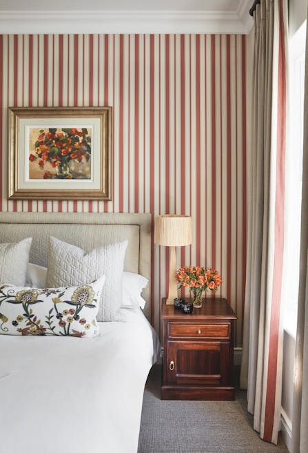 A bedroom with red and white striped wallpaper, a table lamp on wooden bedside table and an artwork of a vase of flowers hangs above the bed.