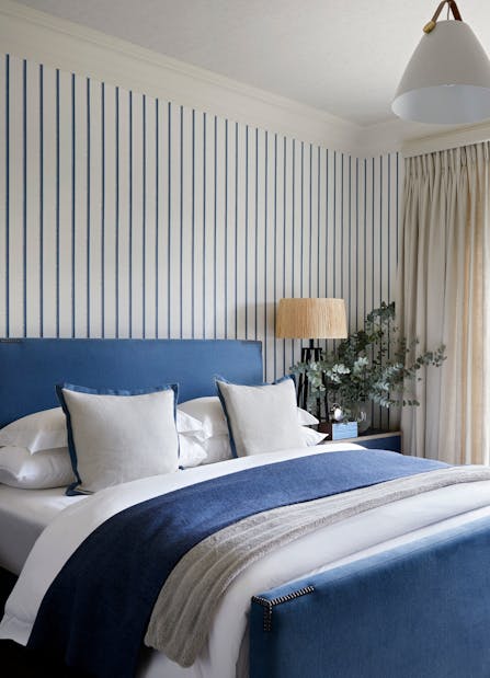 A contemporary bedroom with blue and white striped wallpaper, and a navy blue bed layered in blue with white and grey bedding.
