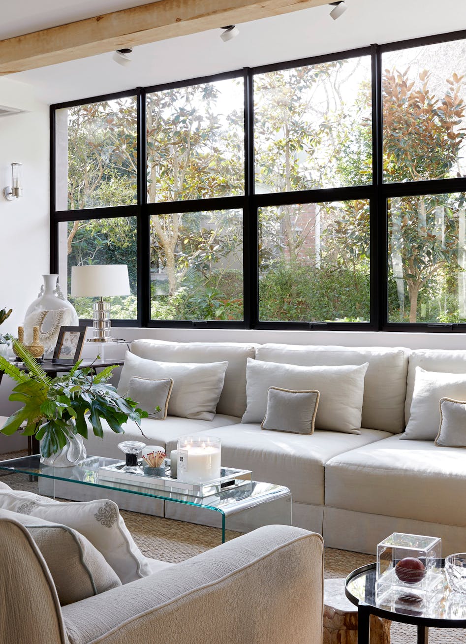 A modern living room with a glass coffee table, a white sofa, and large windows with a view of the greenery outside.