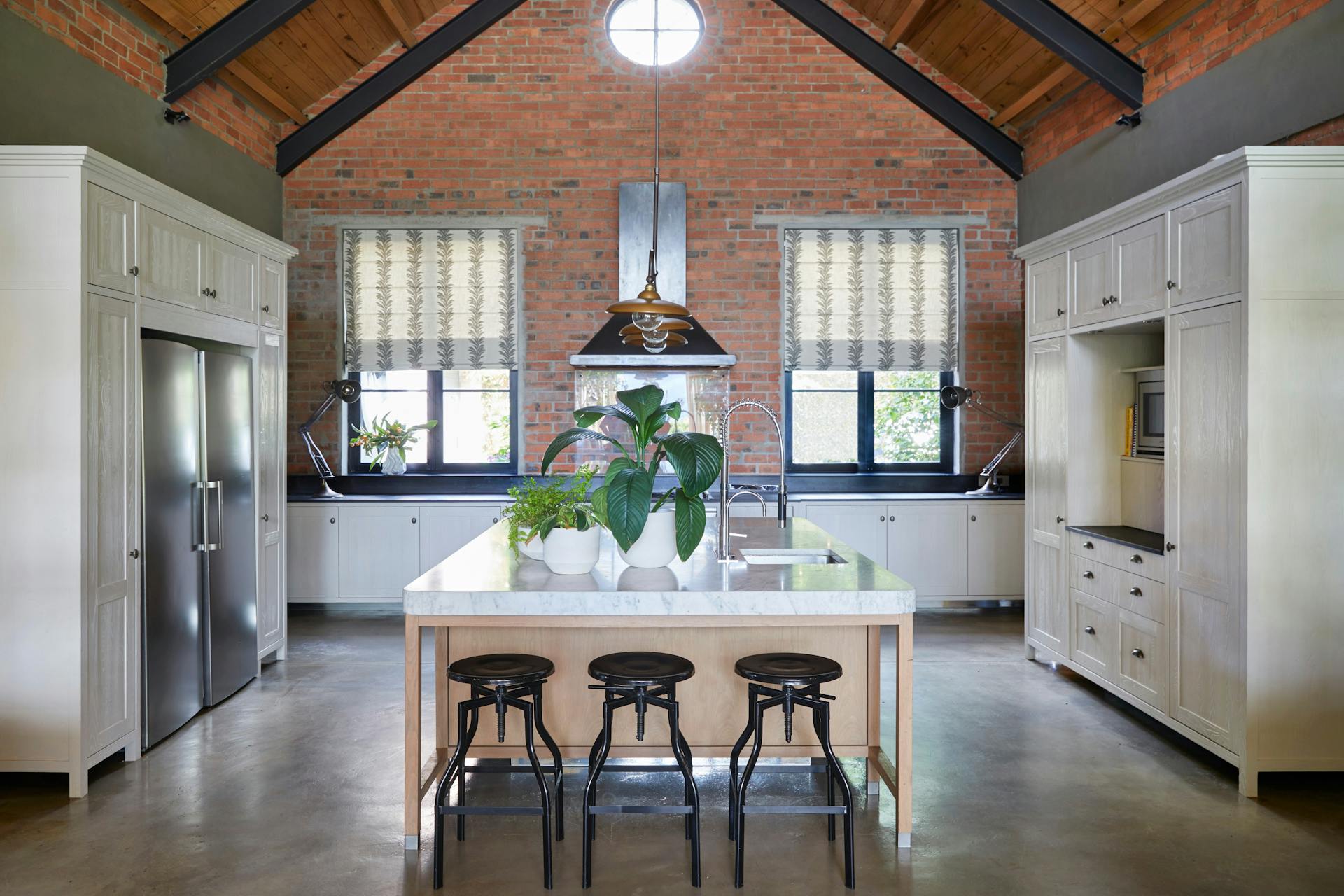A modern, A-frame kitchen with an exposed brick wall features a central kitchen island and white cabinetry. Bar stools are arranged in front of the island and potted plants sit on its surface