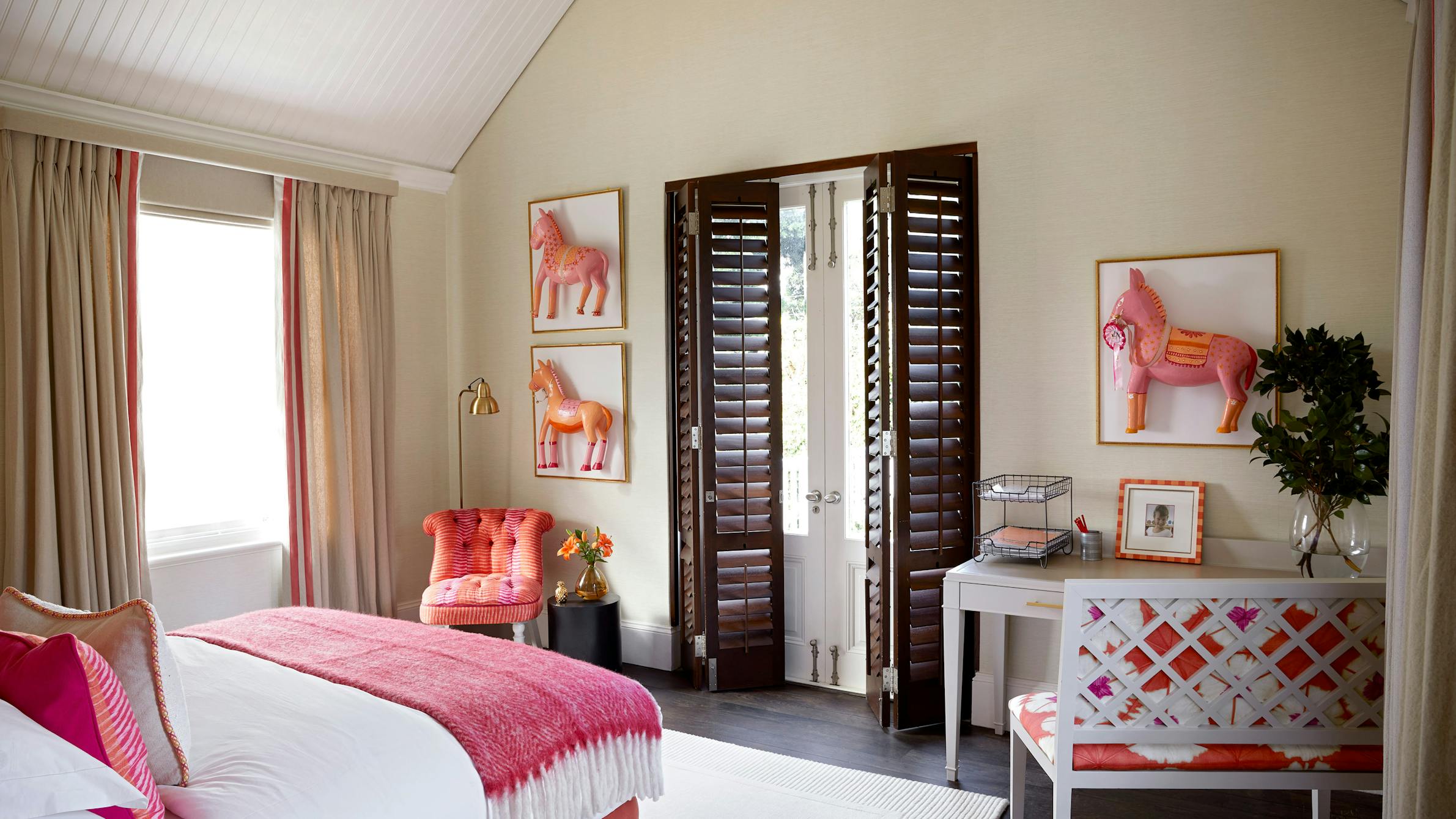 A children's bedroom that features a bed with pink pillows and bedding, a pink occasional in the corner, wooden shutters and a desk with a chair to the right.