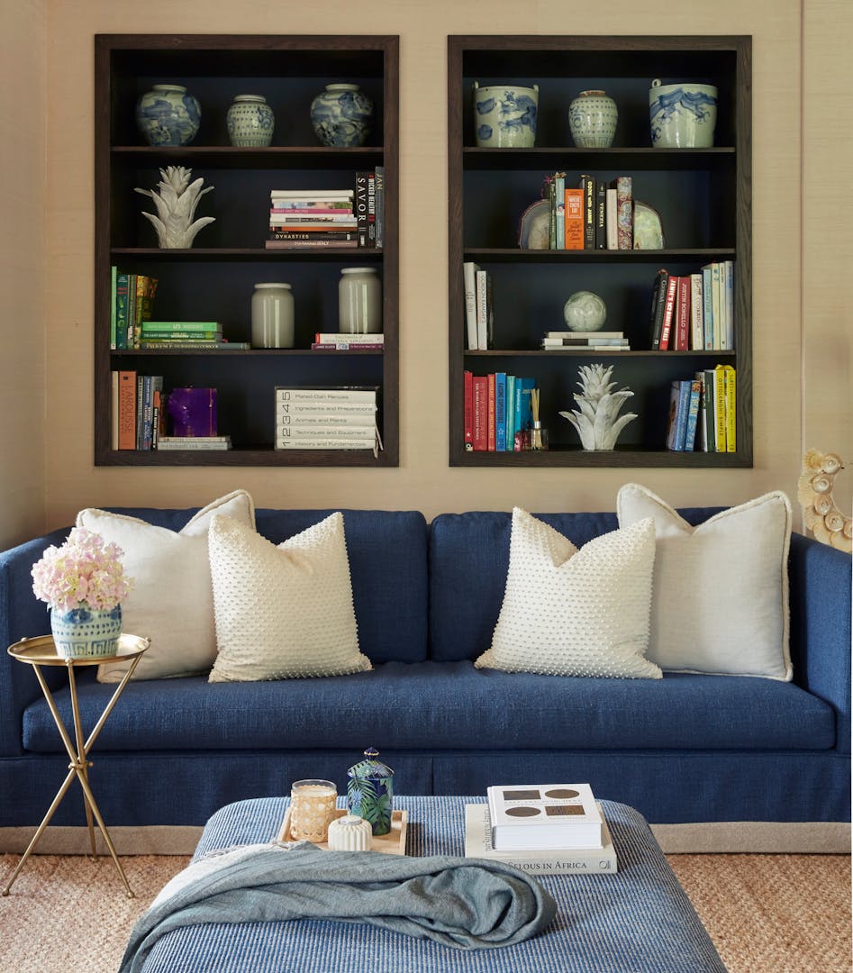 A large, dark blue sofa holds four white cushions. It sits in front of a bookshelf holding cookbooks, art publications and a variety of decorative ceramic pieces. In the foreground, a blanket, two art books and a tray holding three decorative ornaments sit on a square ottoman. A small metal side table holds a vase of flowers.