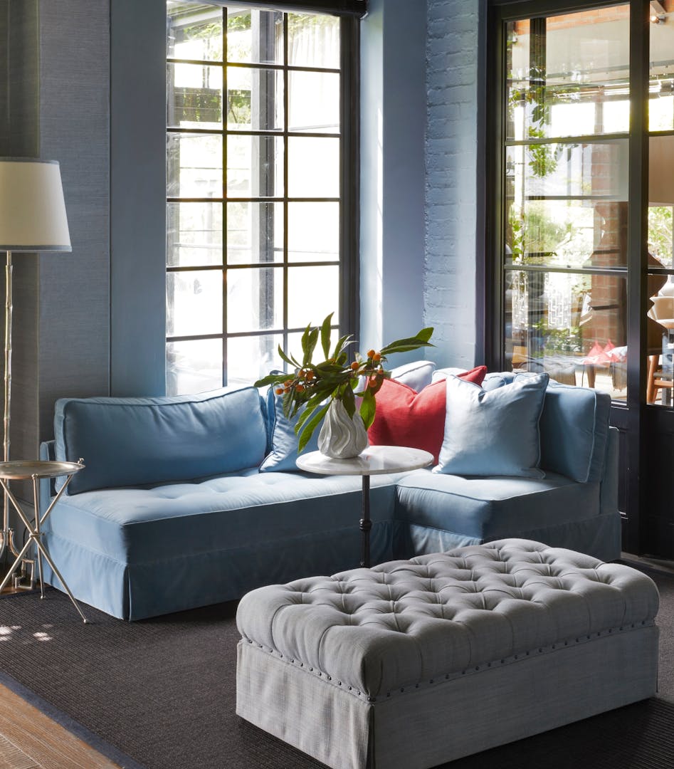 A blue corner chaise enjoys light from windows on two of its sides, while a grey-upholstered ottoman with button detailing is in the centre of the image. A slim, white-topped table holds a vase filled with leaves and small fruit