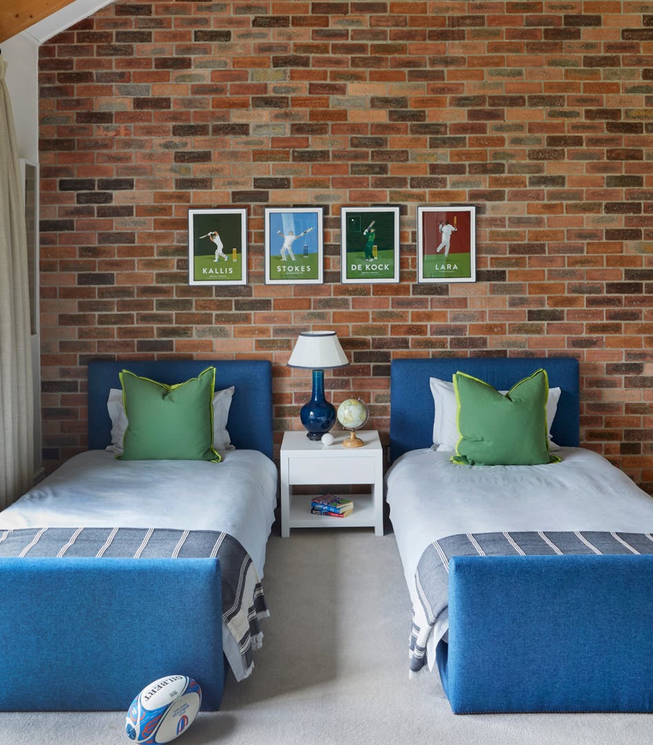 Twin beds with blue and white linen are separated by a small, white bedside table holding a blue lamp with a white shade, a globe, and some books. Behind the bed, the exposed-brick wall displays four framed, simplified images of famous cricketers and the words "Kallis", "Stokes", "De Kock" and "Lara" respectively. A Gilbert rugby ball is on the floor