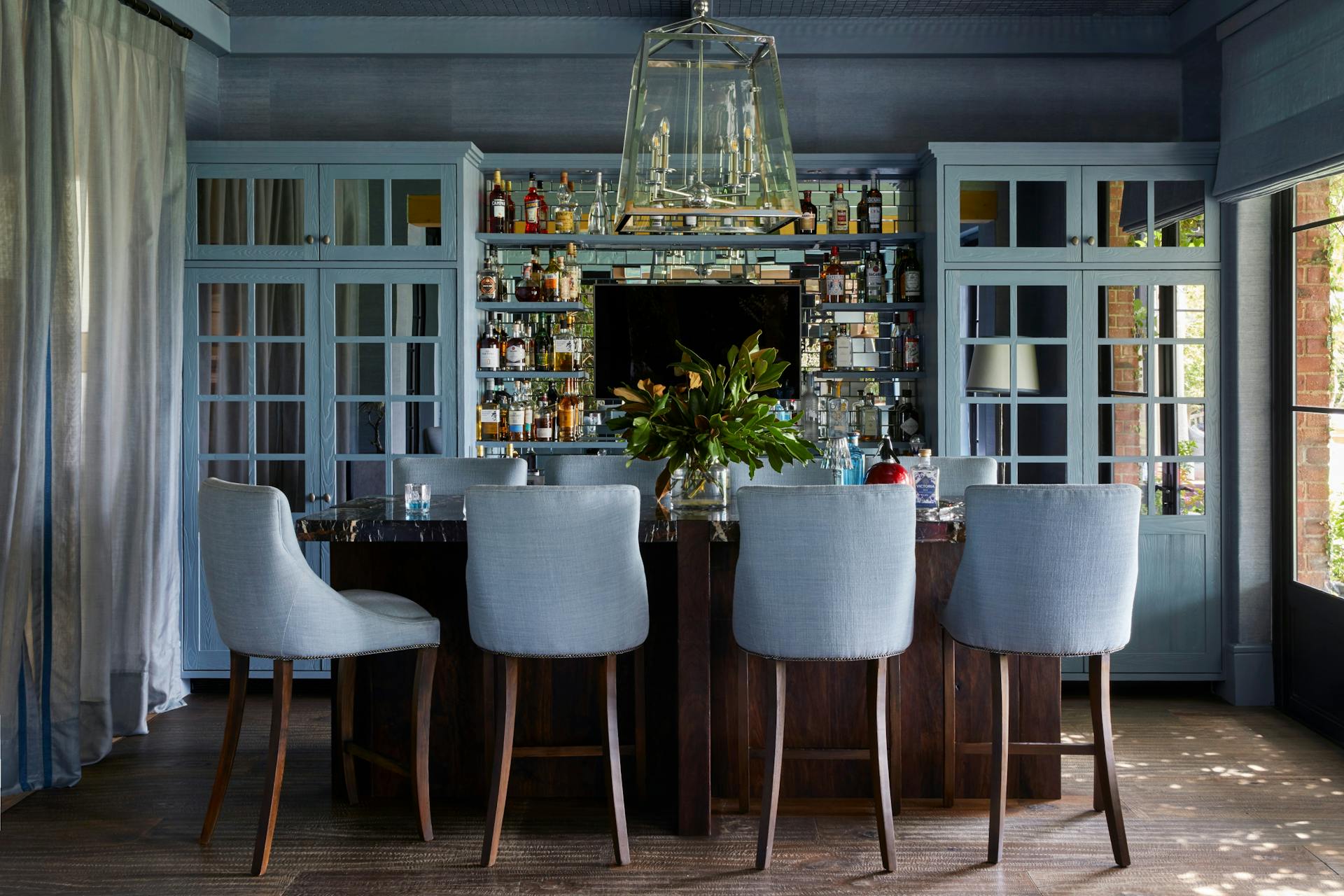 Four blue-upholstered bar chairs sit in front of a wooden bar counter holding a large glass vase of greenery. Under a central, angular hanging light fitting, the bar walls accommodates four shelves of liquor bottles, with large, glass-paned doors to the left and right