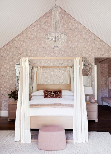 A sophisticated bedroom with a 4-poster bed in the centre, patterned wallpaper on the back wall and a pouffe just in front of the bed on the rug.