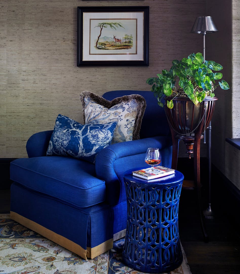 A blue armchair featuring two richly patterned cushions sits on a patterned rug with a ceramic, cut-out side table next to it. On the wall is a framed illustration of an antelope, and slightly behind the chair are a leafy plant and a metallic standing lamp