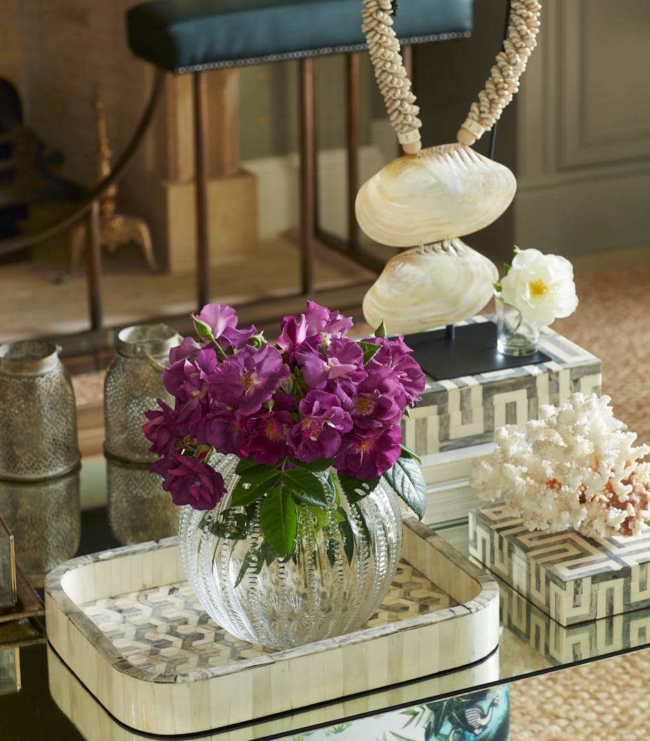 A closeup of a coffee table shows various trays and boxes adorned with geometric patterns, holding ornaments, coral, and a glass vase of flowers.