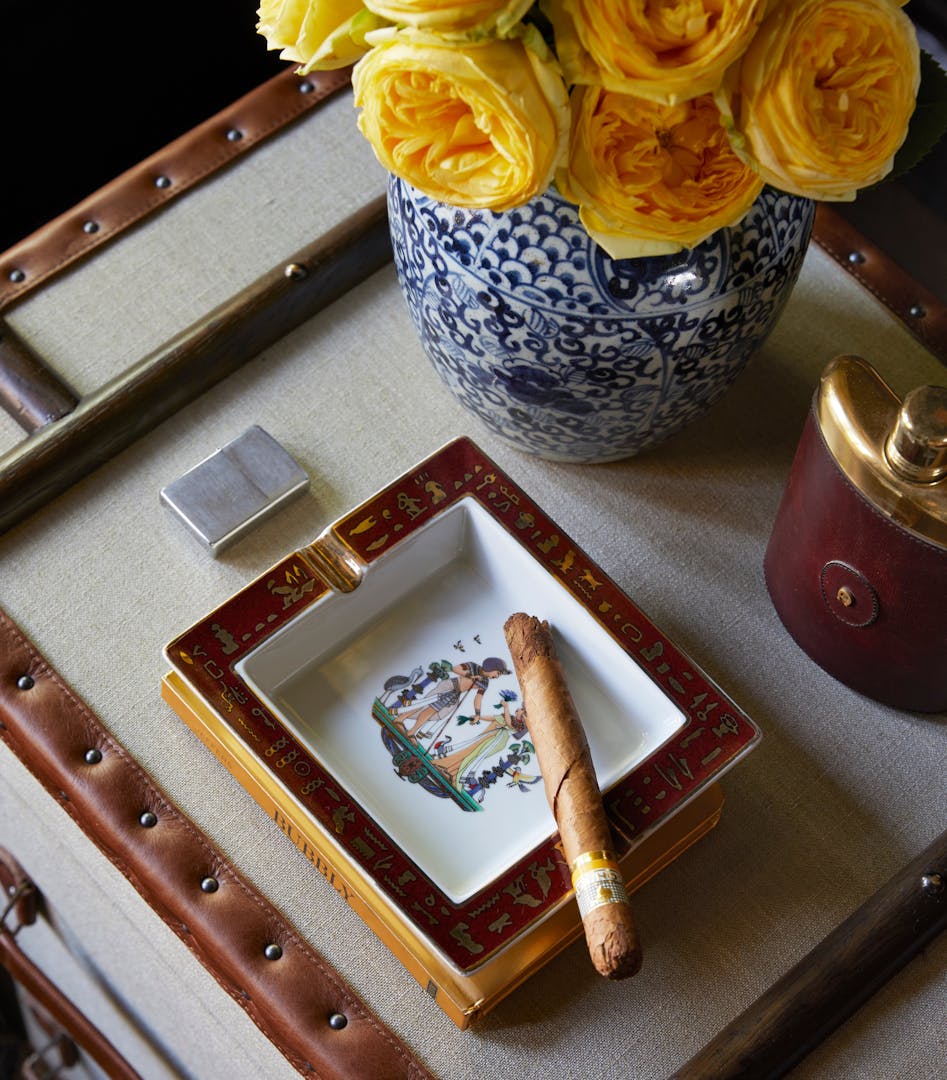 A close-up of a cigar in a square ashtray with a red frame, metallic details and a central image that looks like Egyptian art. Around the ashtray are a hip flask, Zippo lighter and a blue-and-white vase holding yellow flowers