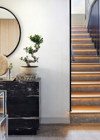 A console table with a bonsai plant atop and a circular mirror on the wall next to a staircase leading upstairs.