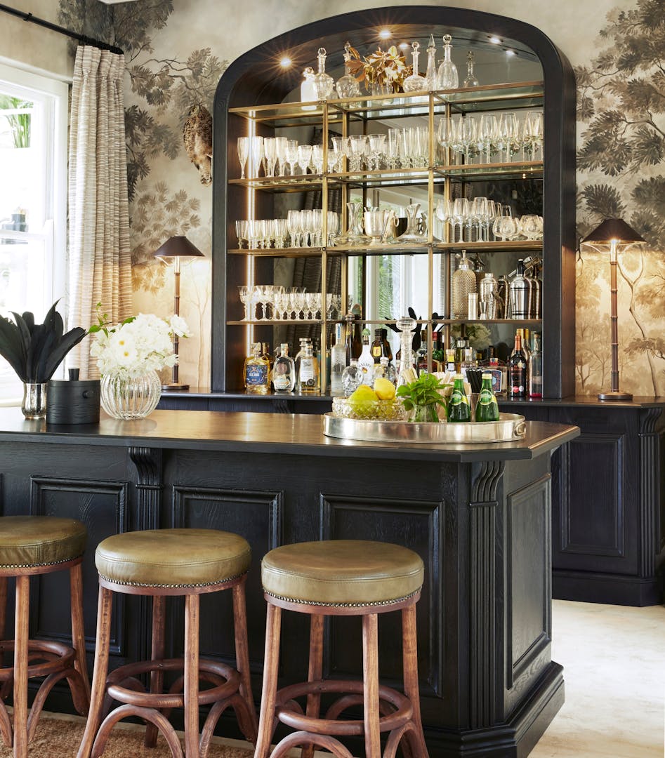 A dark wood bar with three leather-seated bar stools stands in front of a curved, mirrored sideboard holding stemware, decanters and bottles of alcohol.