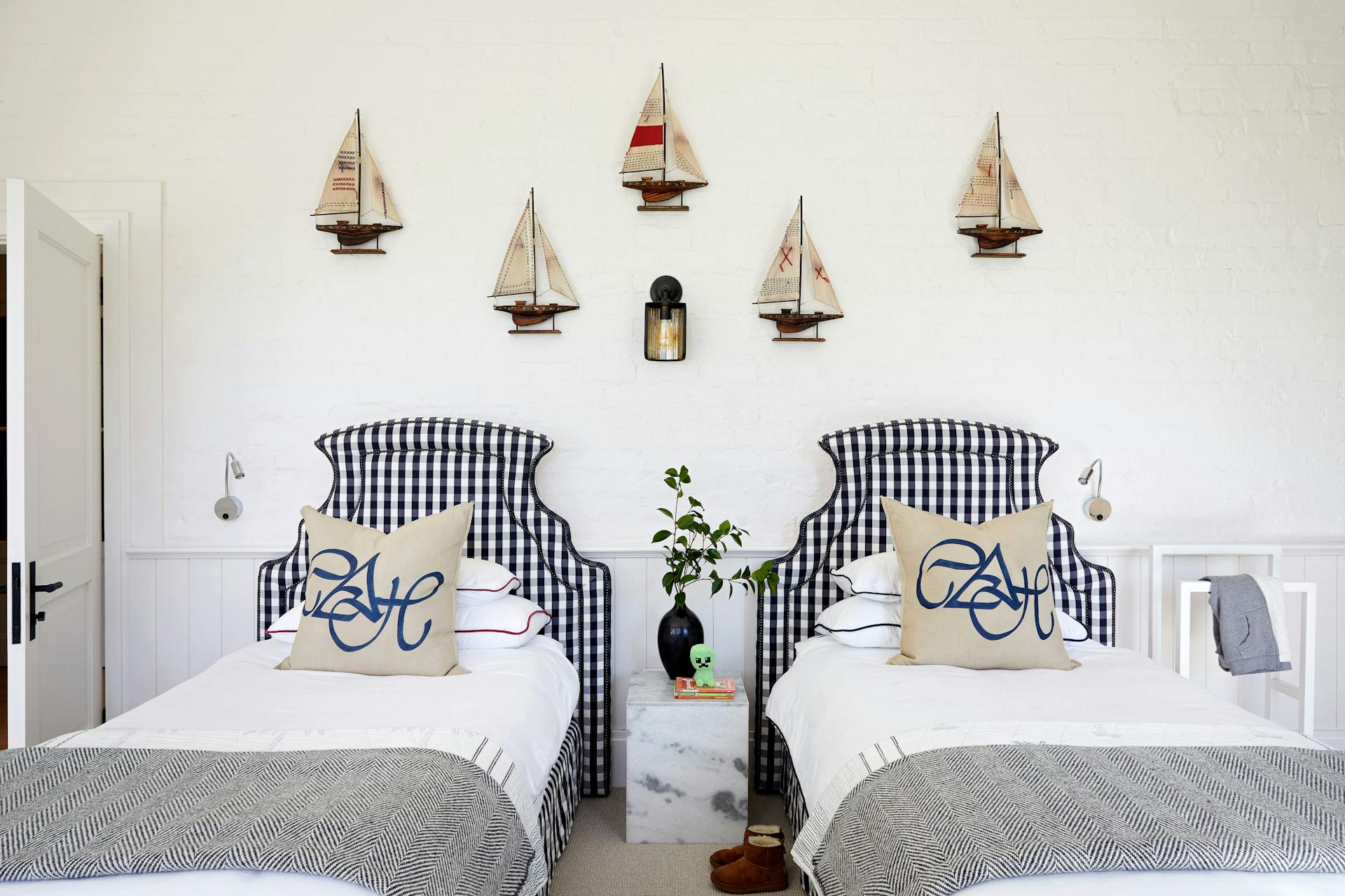 A marine-inspired bedroom with two single beds that both feature blue and white checkered headboards. Artwork of ships hang on the wall with a side table in between the beds.