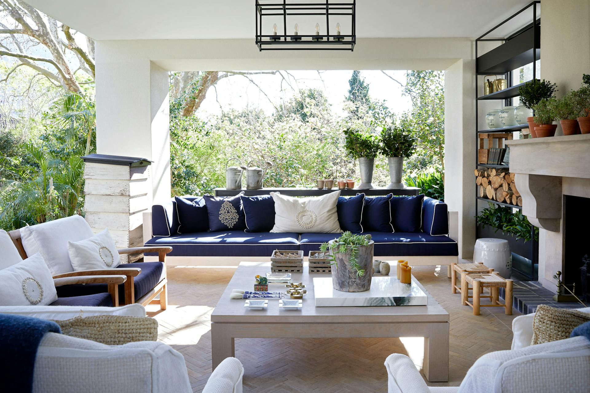An outdoor entertainment area on a patio with a square coffee table in the foreground, a deep blue sofa behind it, and lush greenery in the background.