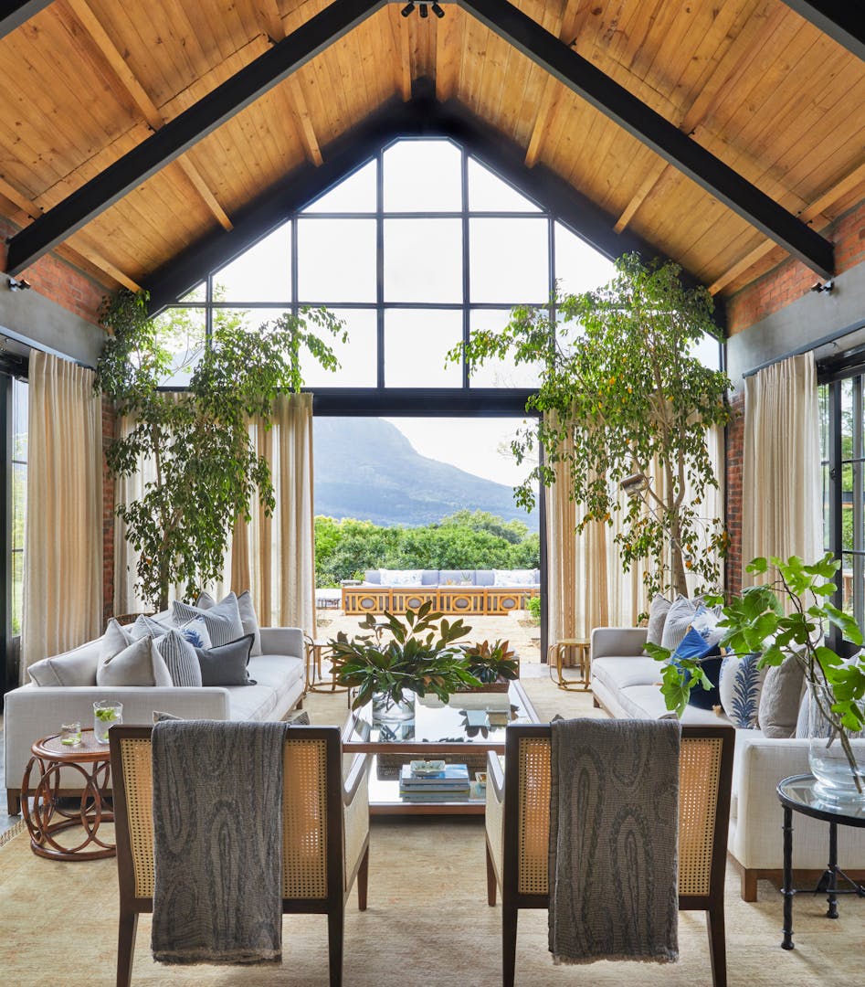 A living room with an a-frame roof, two occasional chairs and two sofas in the foreground with large plants that frame the door to the outside area in the background