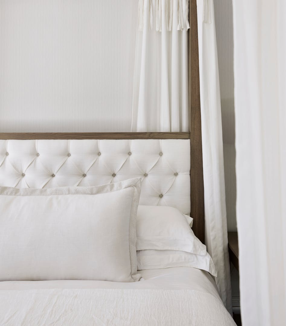 A close-up of a four-poster bed's buttoned headboard upholstered in linen with pillows and duvet in very light-coloured fabrics.