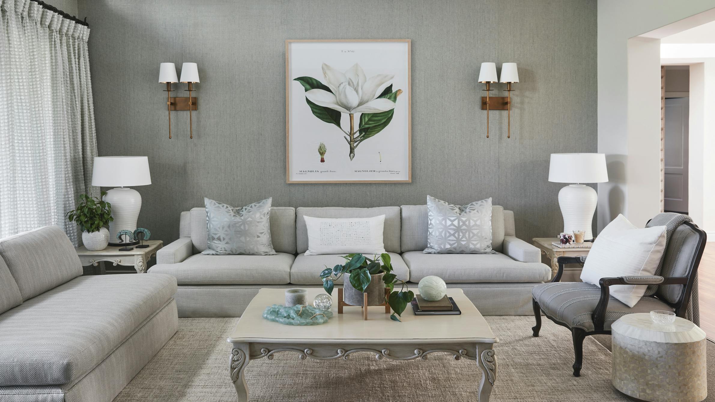 An elegant living room with a neutral, grey colour palette, an ornate coffee table in the middle, surrounded by grey sofas and an armchair with flower artwork on the wall at the back.
