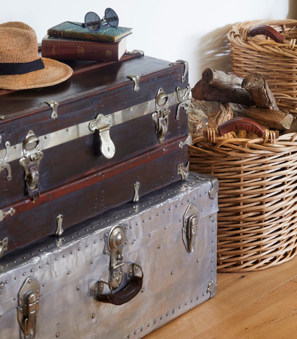 Two old-fashioned luggage chests, one on top of the other, sit next to wicker baskets of fire wood. On the top chest are a panama-style hat, books, and glasses with round frames