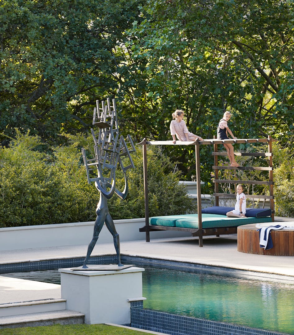 A contemporary sculpture beside an outdoor swimming pool in the foreground and children seated on a 4-poster daybed in the background.