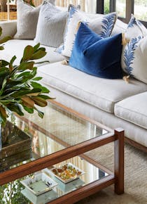 A wooden-framed coffee table with a mirrored base and clear glass table top holds ornate ashtrays on its lower levels and what appears to be a vase of greenery on its top. A neutral coloured couch is visible, holding blue and blue-patterned cushions.