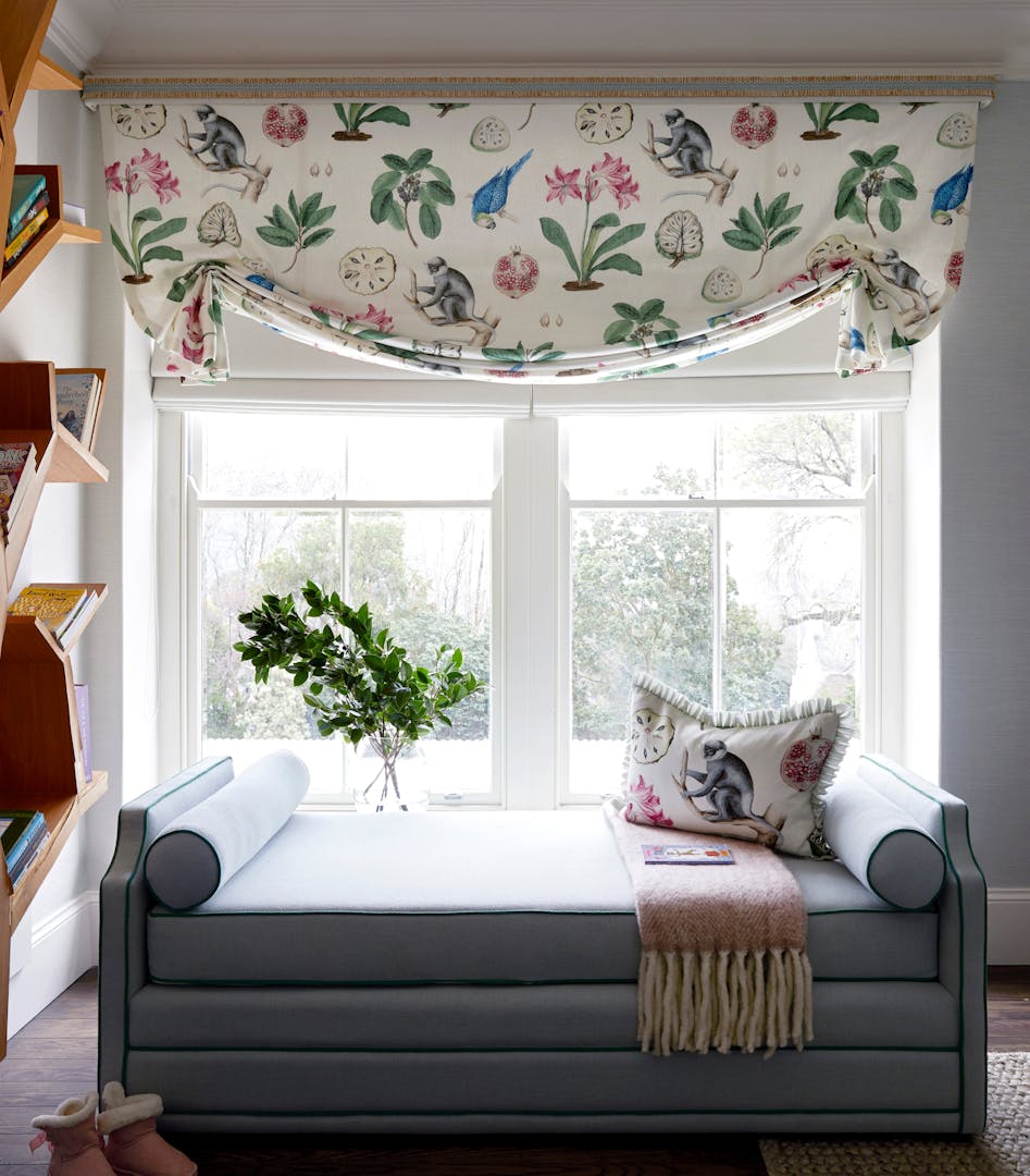 A blue-upholstered chaise sits in front of a window looking onto a garden. On the windowsill is a glass vase holding branches of greenery, and the couch holds a fringed blanket, a magazine, and a cushion made of a fabric depicting monkeys, fruit and botanicals. The window is draped with the same fabric.