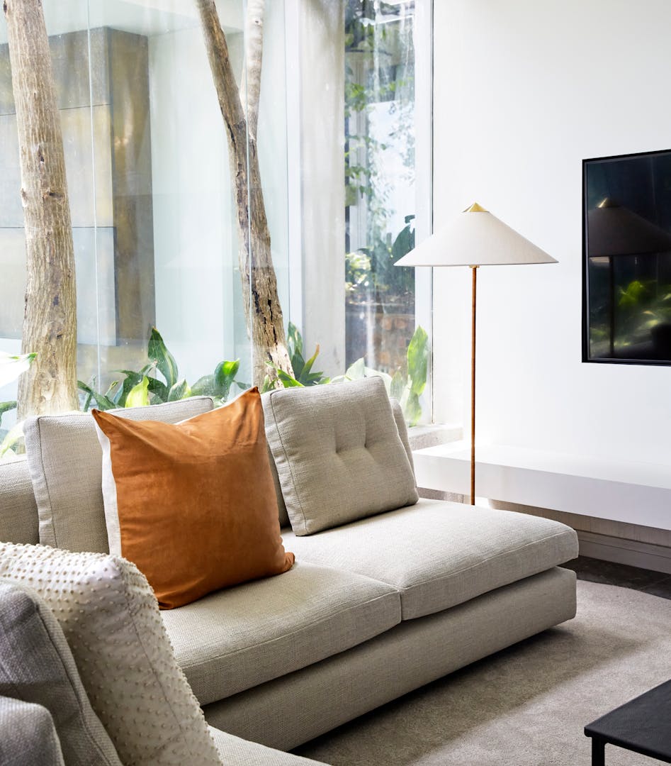 A sofa with a modern floor lamp behind it with glass windows in the background looking onto two trees.