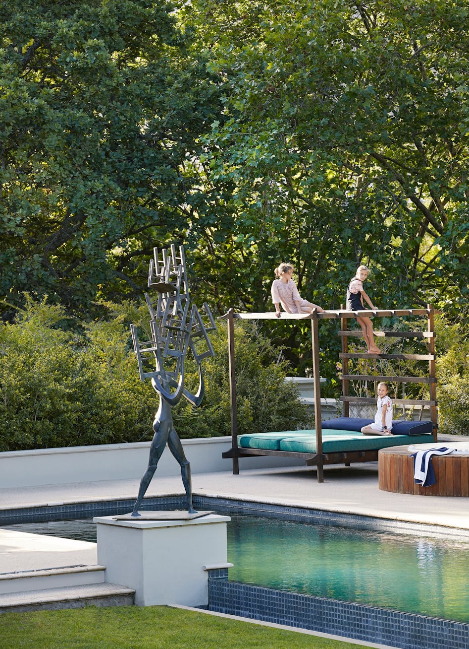 A contemporary sculpture beside an outdoor swimming pool in the foreground and children seated on a 4-poster daybed in the background.