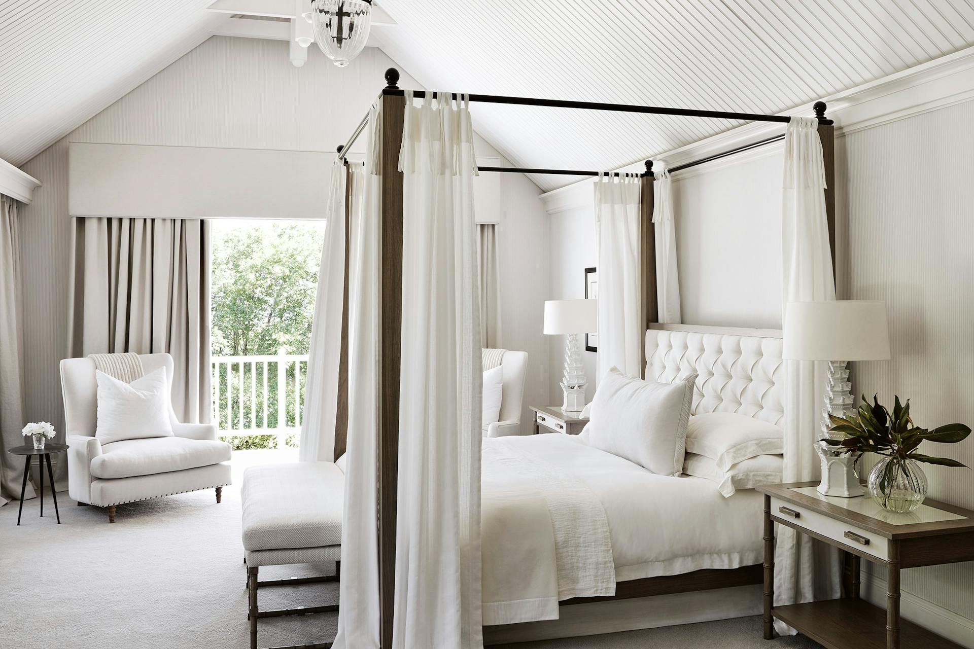 A four-poster bed, upholstered chair, bedroom bench and side table are seen in a bedroom with an A-frame ceiling, furnished in white, light neutrals and dark wood.