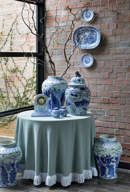 A selection of blue-and-white patterns and illustrated vessels and ornaments sit on and next to a table covered in a green, fringed cloth. On the exposed brick wall behind it, three plates hang, all of them with a design similar to that of the vessels on and around the table