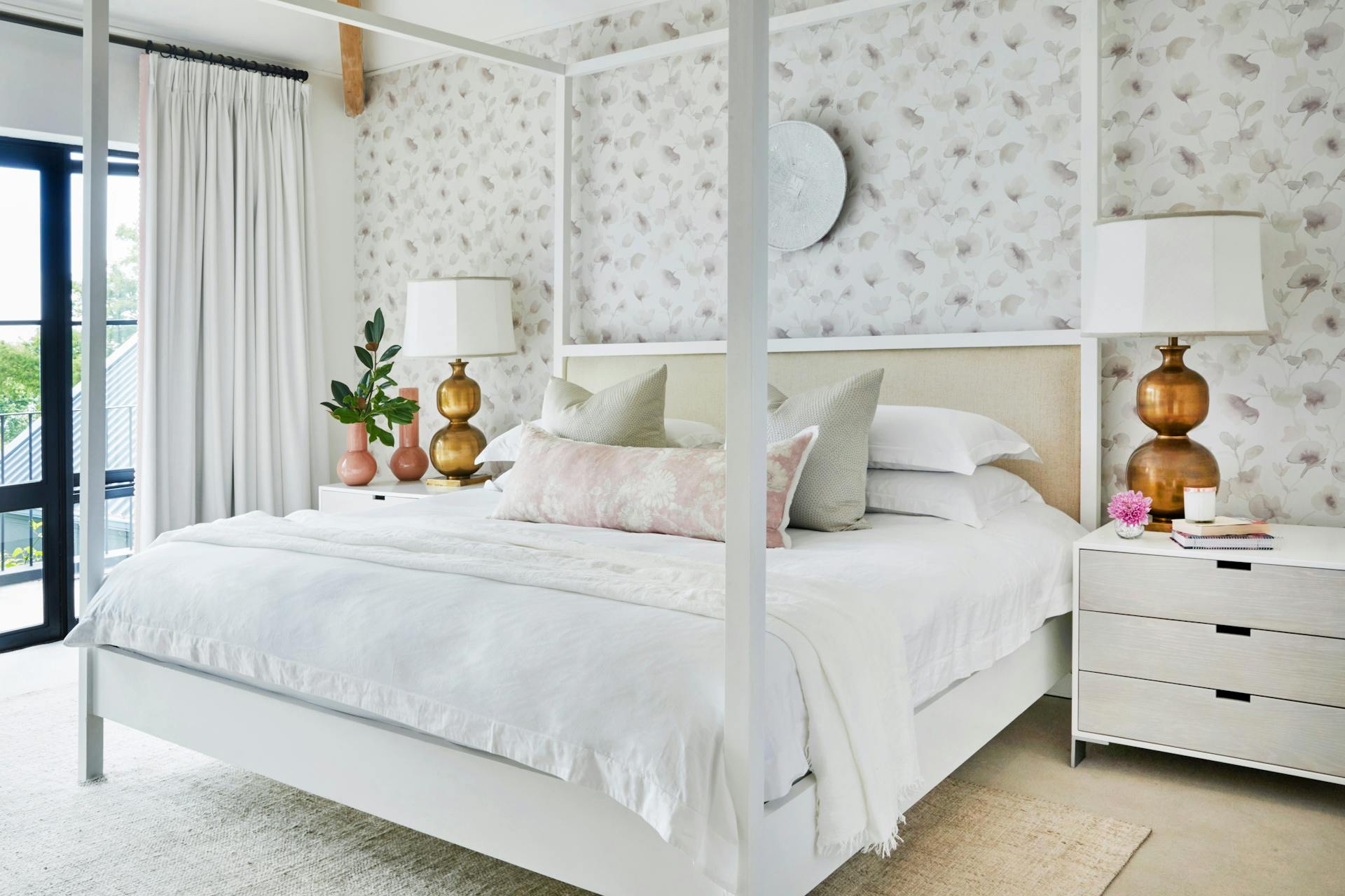 A wide four-poster bed with a white frame, white linen and pale-coloured pillows sits in front of a wall papered in a pale, sepia botanical print. White side tables to the left and right of the bed hold metallic lamps and vases.