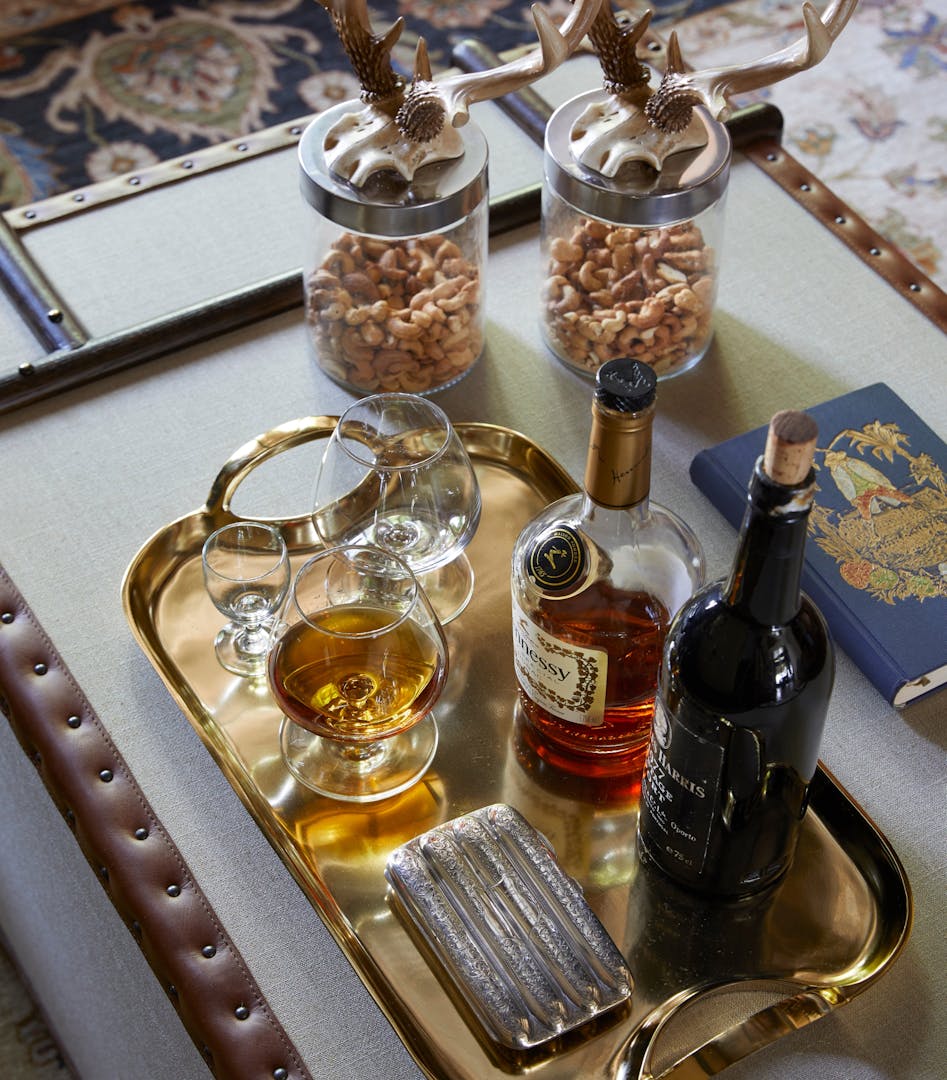 A bottle of Hennessy and a bottle of port sit on a gold tray with some glasses and what appears to be a cigarette box. Two jars contain cashew nuts, and in the background a patterned carpet is visible