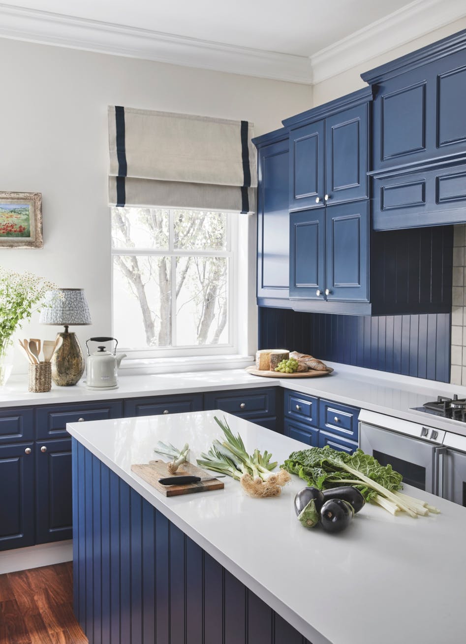 A kitchen with deep blue cabinetry, a window with a view of the trees outside and a kitchen island with vegetables on top.