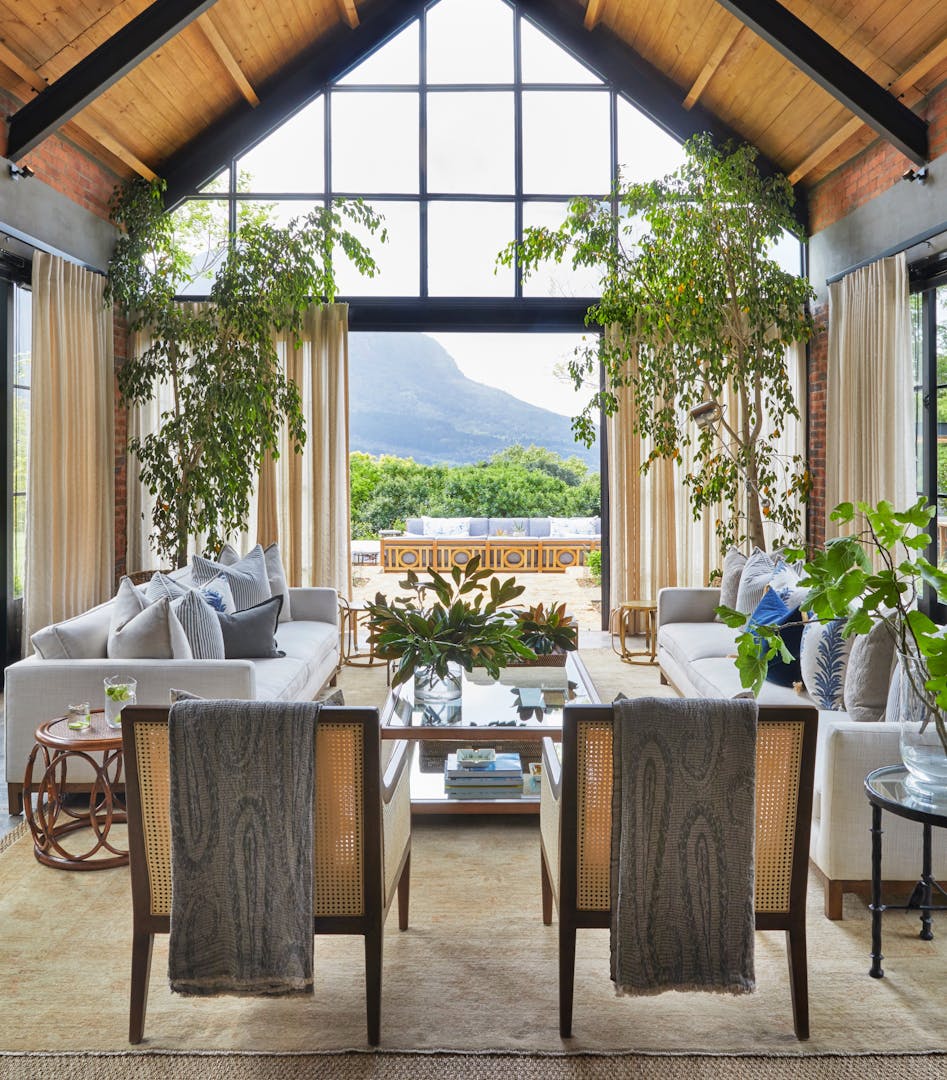 An A-frame, double-volume ceiling frames a living area decorated in neutral tones with natural wood and fresh greenery. An outdoor area and mountain views are visible in the background