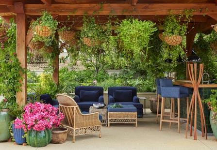 An outdoor entertaining area featuring rattan furniture, surrounded by greenery, with a bar area to the left.