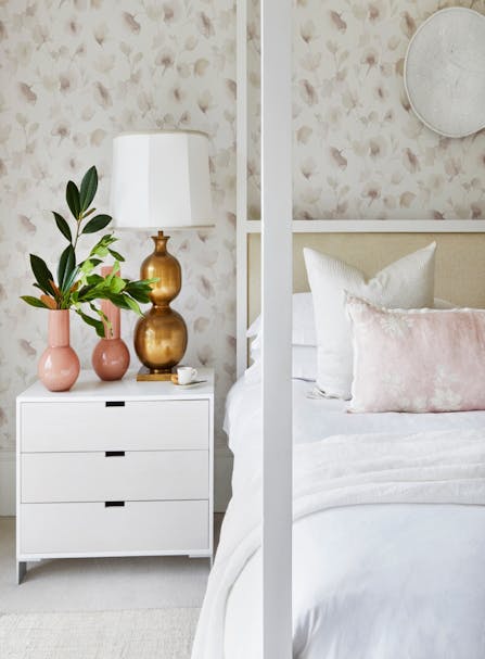 One side of a four-poster bed with white linen is seen beside a set of bedside drawers. On the top of the drawers are a white espresso cup, two metallic vases and a metallic lamp base with a white lampshade. One vase holds a few branches of green leaves, and the wallpaper features a pale pattern of leaves or florals.