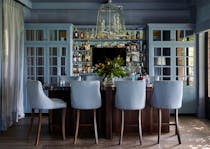 Four blue-upholstered bar chairs sit in front of a wooden bar counter holding a large glass vase of greenery. Under a central, angular hanging light fitting, the bar walls accommodates four shelves of liquor bottles, with large, glass-paned doors to the left and right.