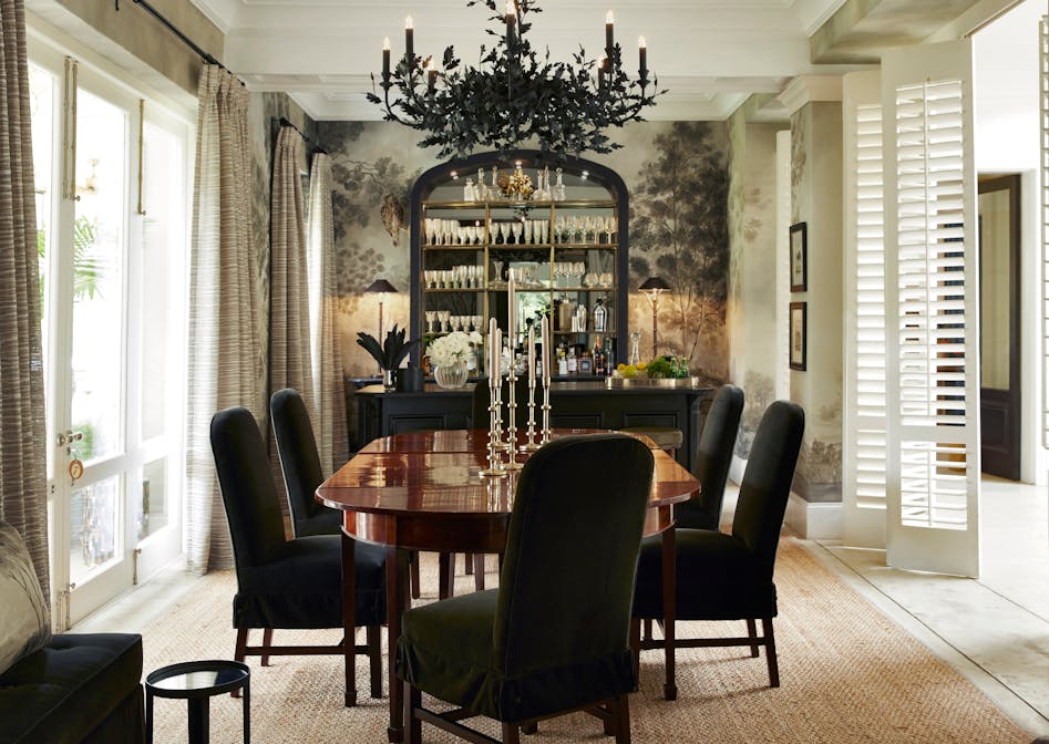 A polished wood dining table holds metal candlesticks and is surrounded by six chairs in a dark fabric. In the background, a tall mirrored sideboard holds stemware, vases and a pair of lamps.