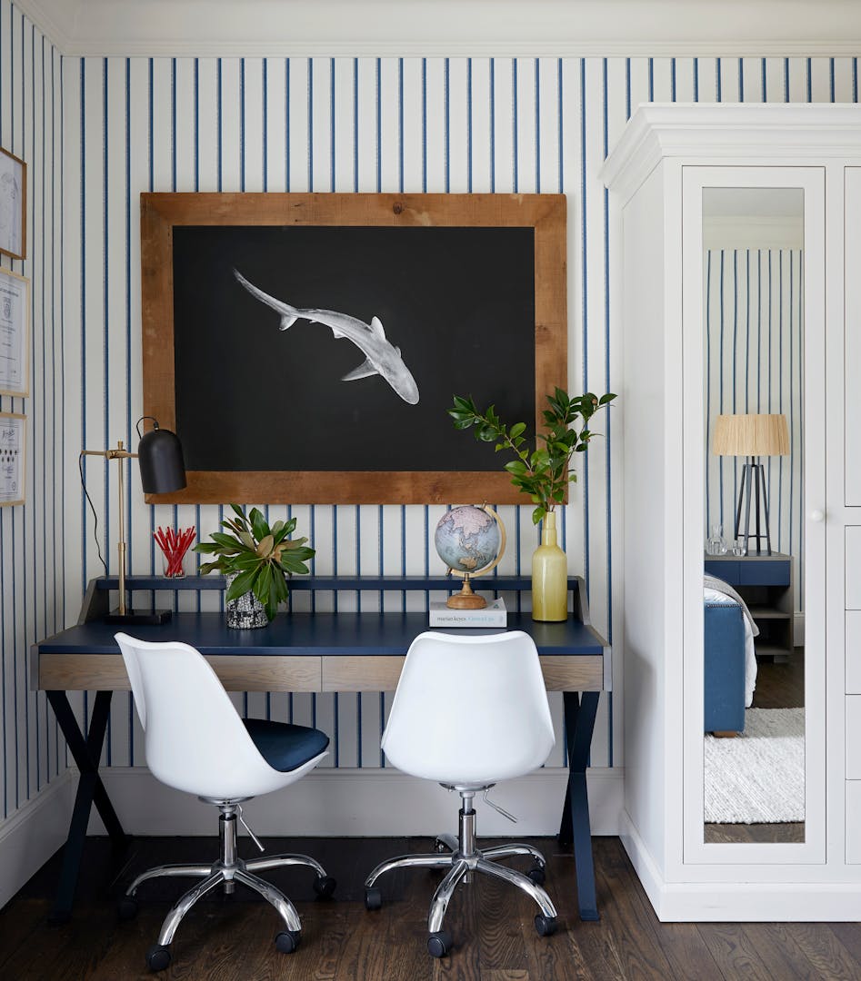 A framed, black-and-white image of a swimming shark or fish hangs above a desk that holds a globe, a jar of pencils, an angle-poise lamp and two vases of greenery. Two white chairs stand in front of the desk, and the wall is papered with blue and white stripes.