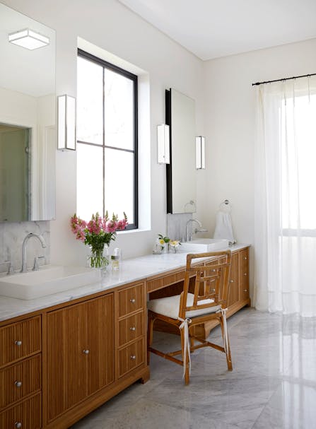 A bathroom counter with two basins on either side and a large window on the wall in the middle. Wooden, slatted cupboards and a wooden chair complete the image.