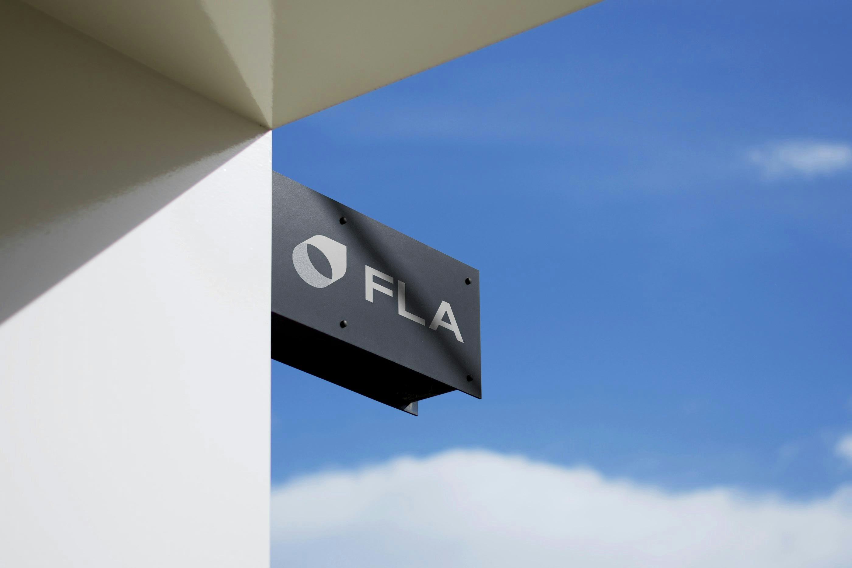 Insegna esterna minimal con logo e scritta 'FLA' montata su edificio bianco, fotografata contro un cielo azzurro. L’immagine rappresenta identità visiva chiara, modernità e riconoscibilità del brand FederlegnoArredo.