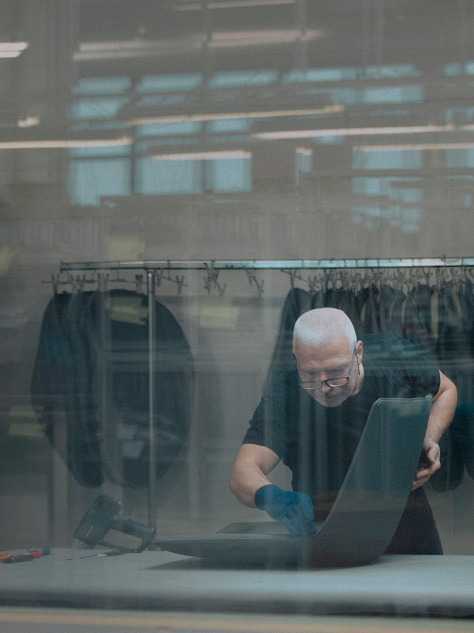 Craftsman working on a modern furniture component inside a wood-furniture sector factory, with clothes hanging in the background. The image conveys artisanal care, innovation, and the value of Made in Italy, aligned with the values of the FederlegnoArredo ecosystem.