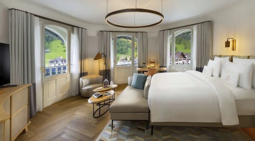 Inside a room at the Kempinski Palace Engelberg holds a beautiful king bed with a white duvet and 3 bright windows showing views of mountains