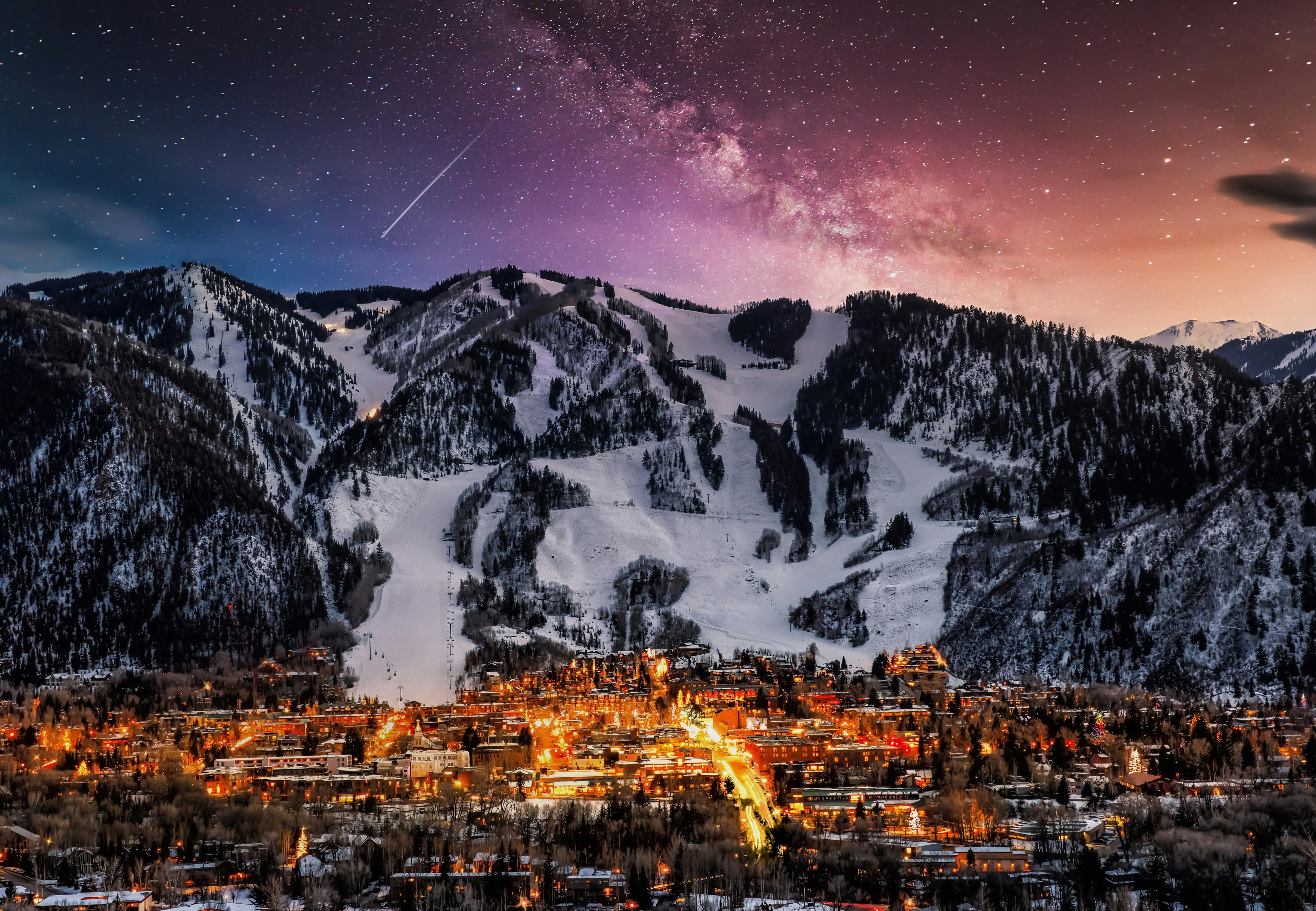 A night view of Aspen Ski Resort, showcasing a city illuminated under a starry sky with majestic mountains in the background.